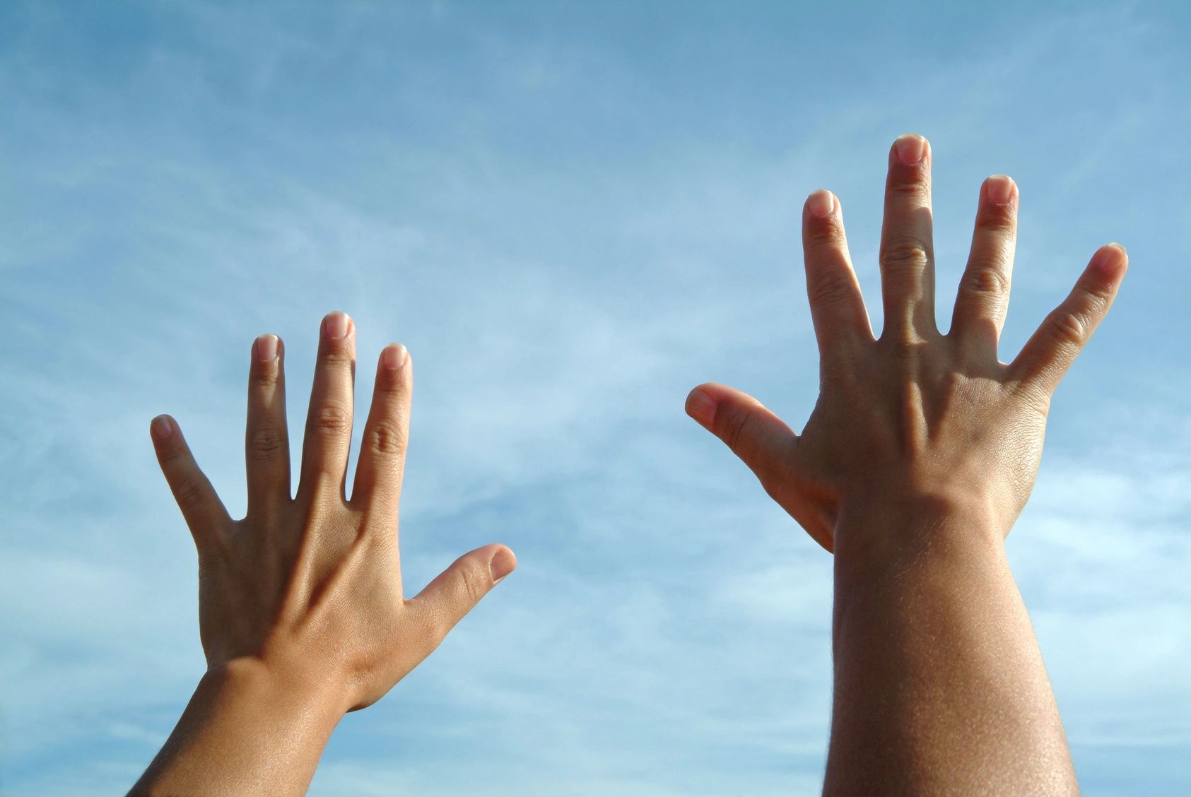 Two hands reaching up into the air in front of a blue sky.